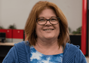 Beyond Vision employee, Mary, wearing a blue sweatshirt while standing and smiling in the office.