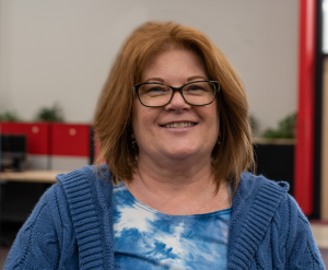 Beyond Vision employee, Mary, wearing a blue sweatshirt while standing and smiling in the office.